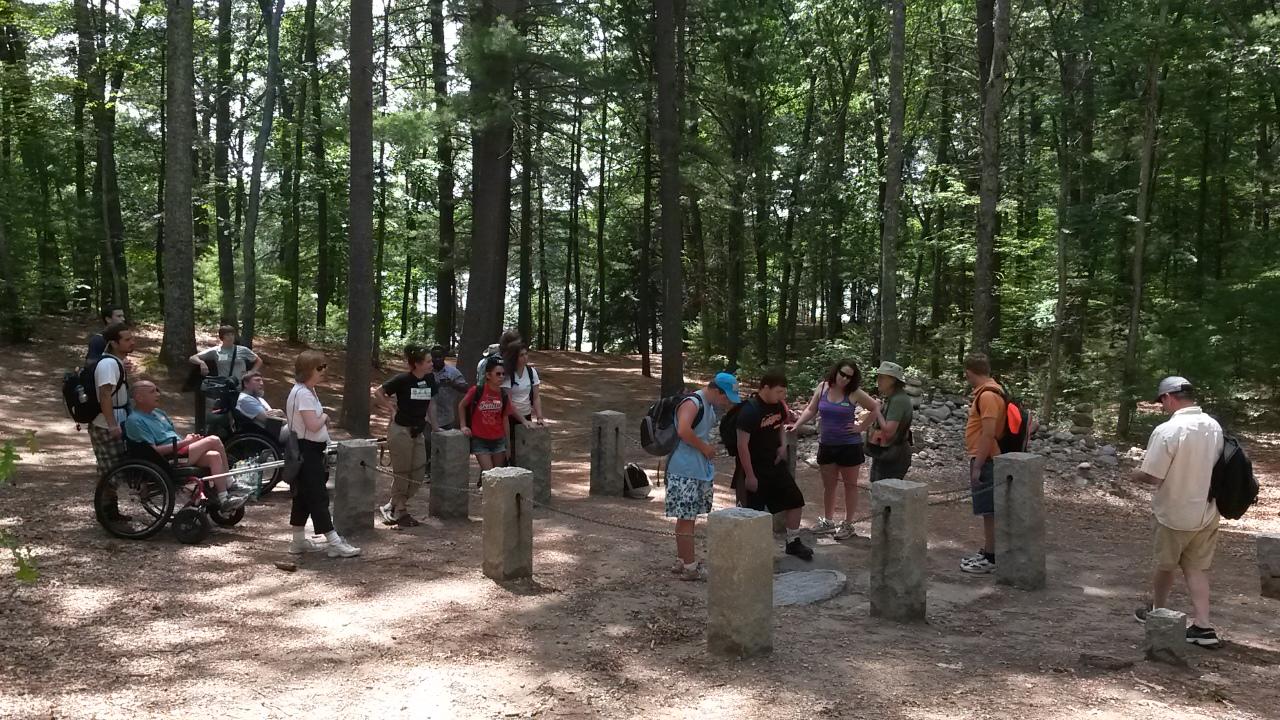 A group of hikers stands in a square of stone pillars in the woods. Some hikers are using hiking wheelchairs.