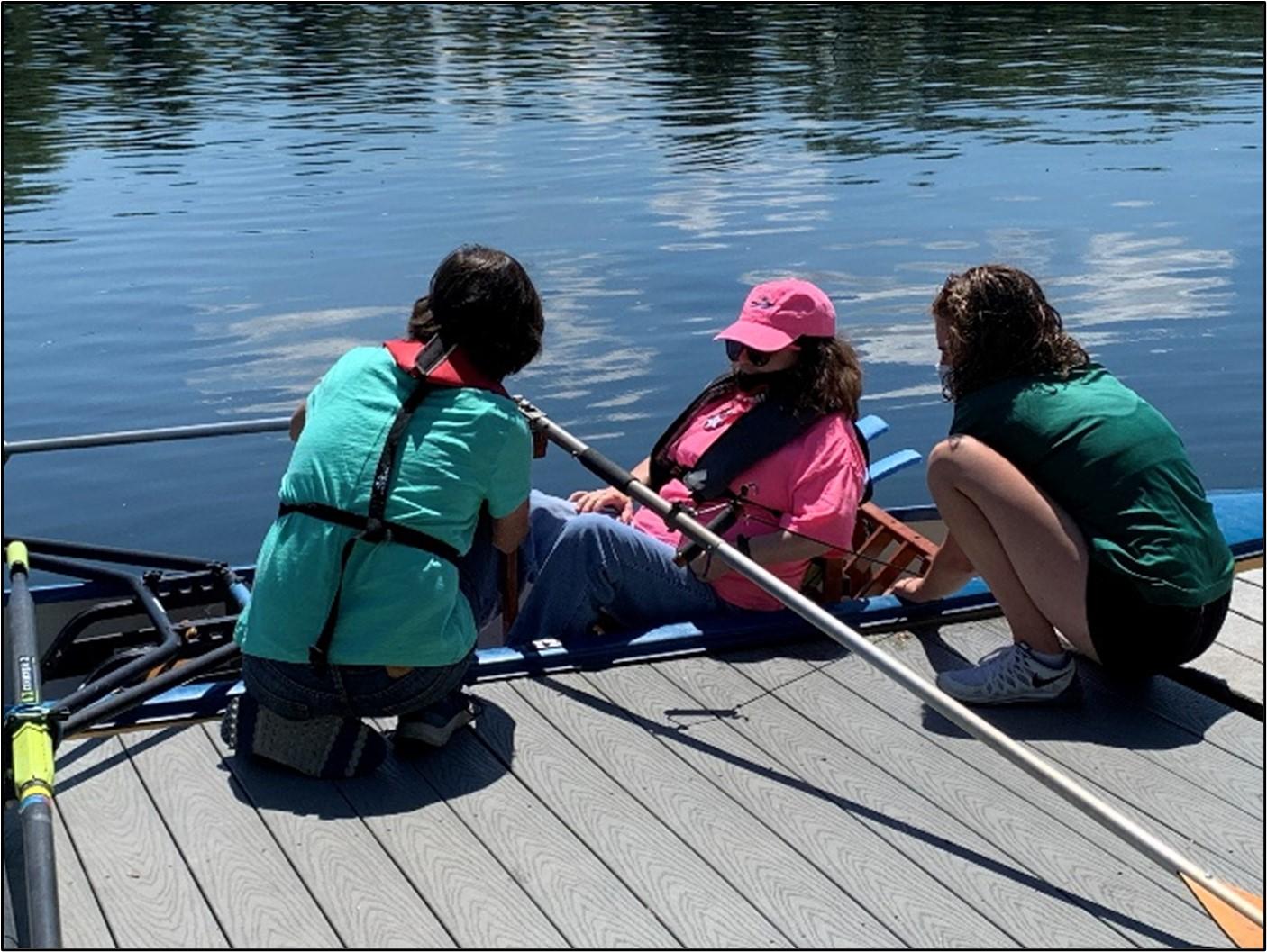 A rower at a dock is assisted by two people. It is a sunny day and the water is calm.