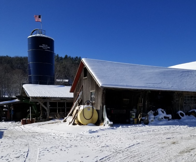 A maple silo with an American flag at the top. The silo is next to a brown farmhouse.