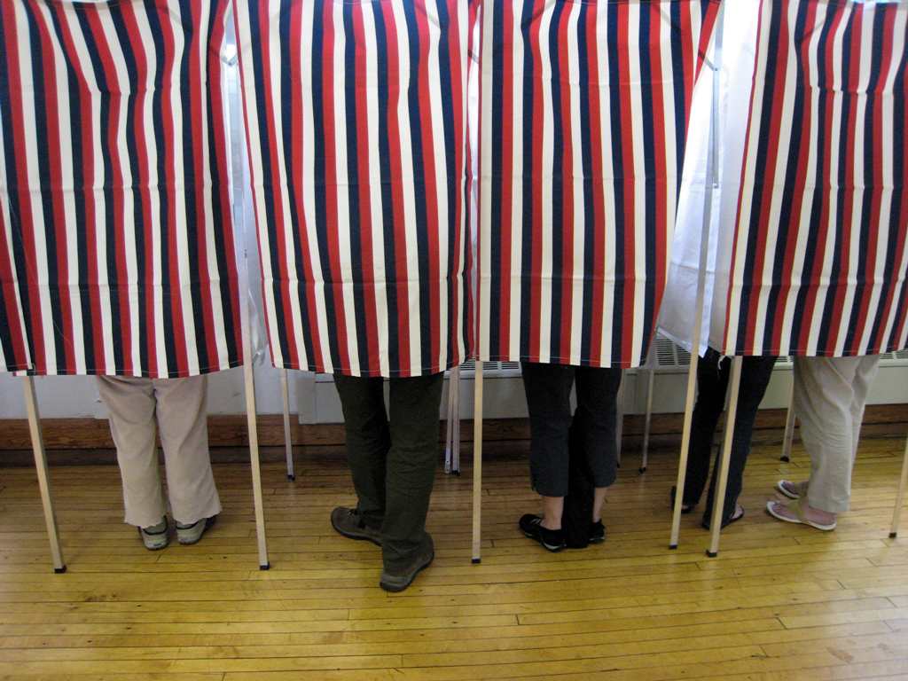 The legs of four people who are hidden by the red and white curtain of a voting booth. 