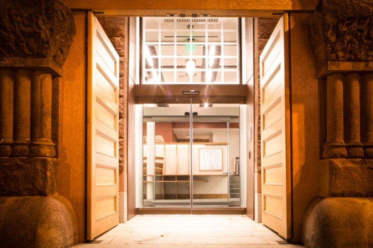 A set of glass double doors recessed into a vestibule with wooden panels that serve as the sides of the vestibule with decorative columns on either side of the vestibule entrance.