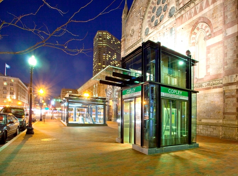 Clear glass elevator entrances on a city street with a building in the background at night