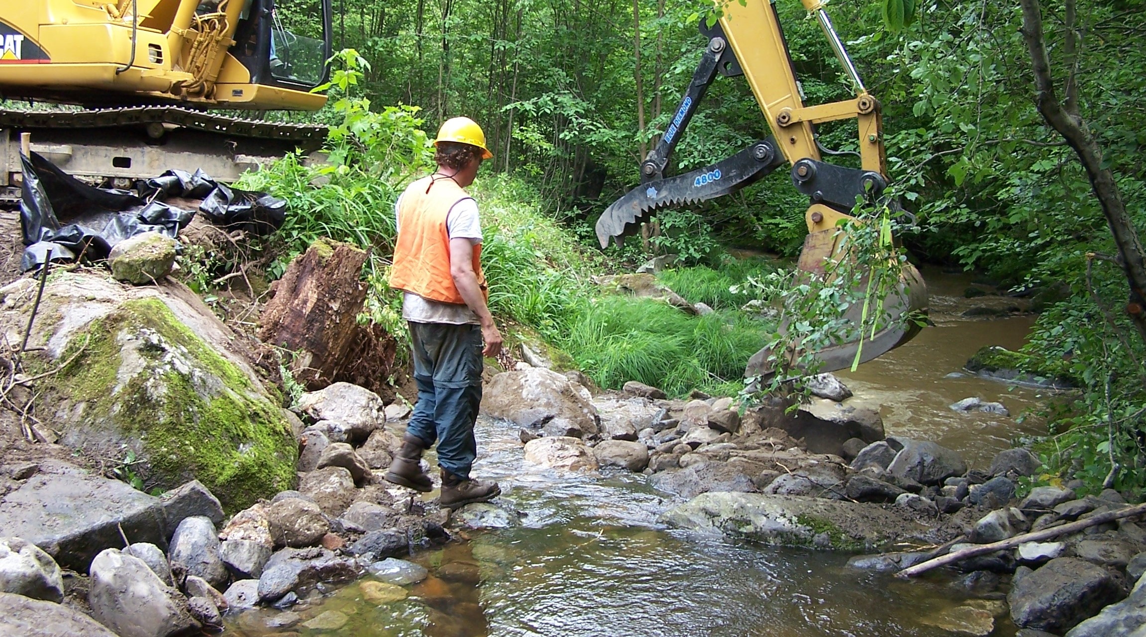 A person in a hard hat and high visibility vest walking in a stream next to construction equipment.