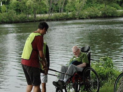 A person wearing a yellow vest is pulling the poles of a hiking wheelchair forward. The person sitting in the hiking wheelchair is smiling and the Charles River is pictured in the background.