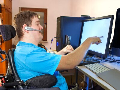 White man in a wheelchair wearing a blue shirt. He has a headset on and is looking and pointing at a computer screen.