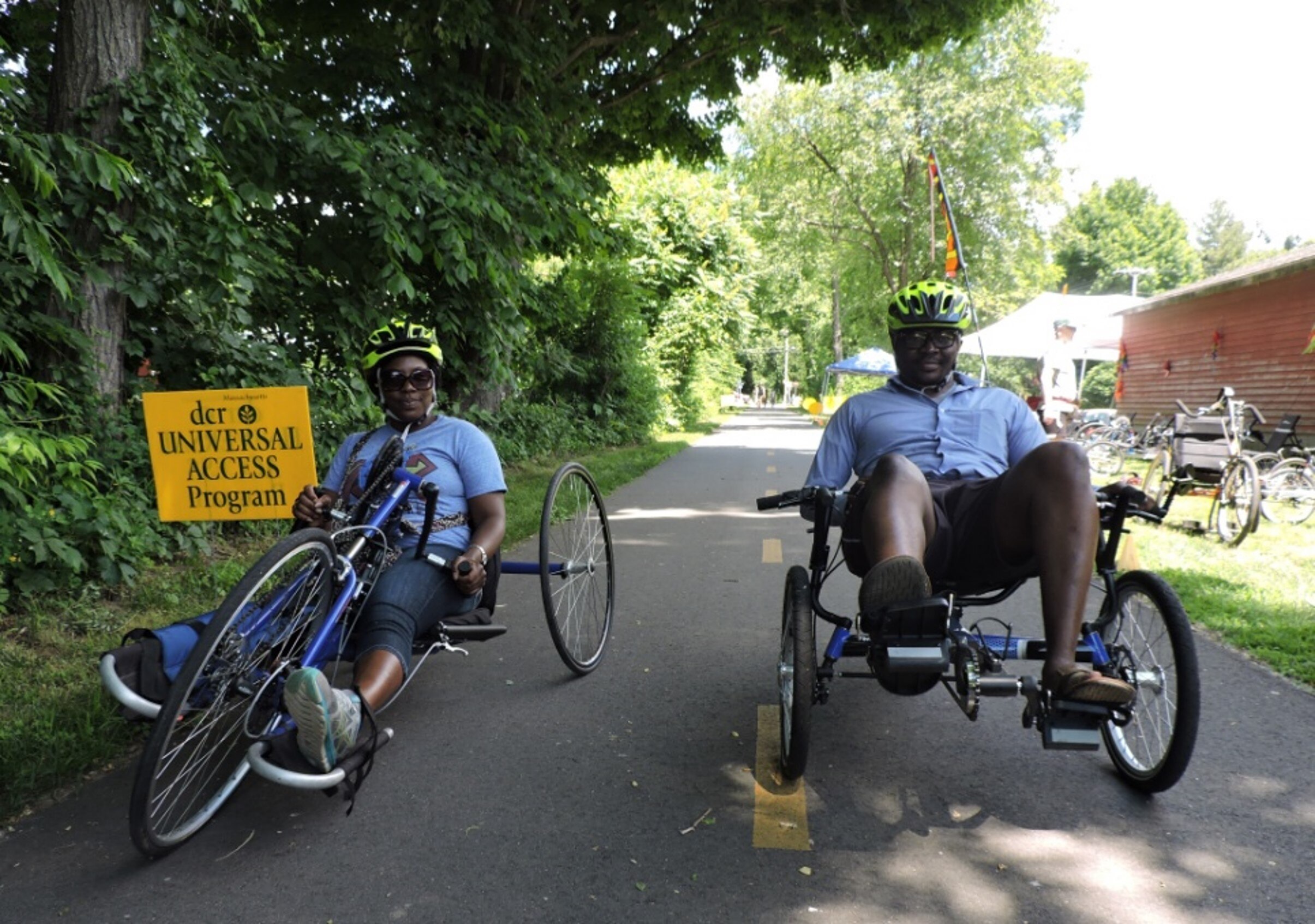 Two cyclers ride side by side on the rail-trail. One using a recumbent cycle the other using a hand cycle. 