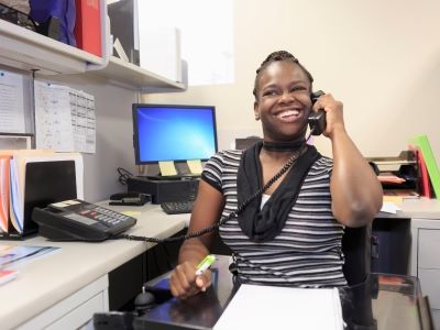 A Black woman using a power wheelchair is sitting at a corner office desk. She is wearing a black and white striped shirt with a black scarf. She is holding a highlighter in her right hand and a desk phone in her right hand, held up to her ear. She has a big smile on her face. The desk is filled with paper files and a desktop computer. 