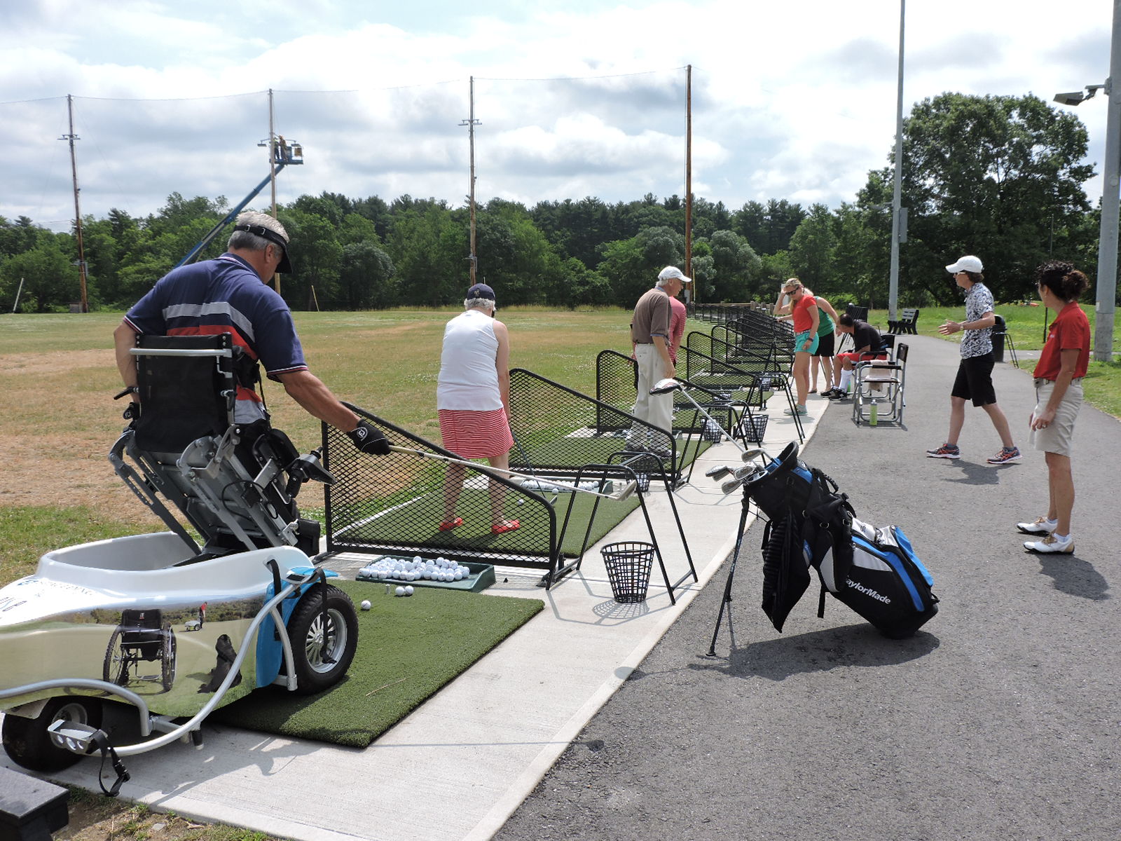 A group of people is standing on the driving range, driving golf balls. Several other people are standing behind them, watching and offering advice. One of the golfers is using a ParaGolfer to hold him upright while he swings. The reflection of a wheelchair, a service dog, and another ParaGolfer user are visible in the side of the ParaGolfer.