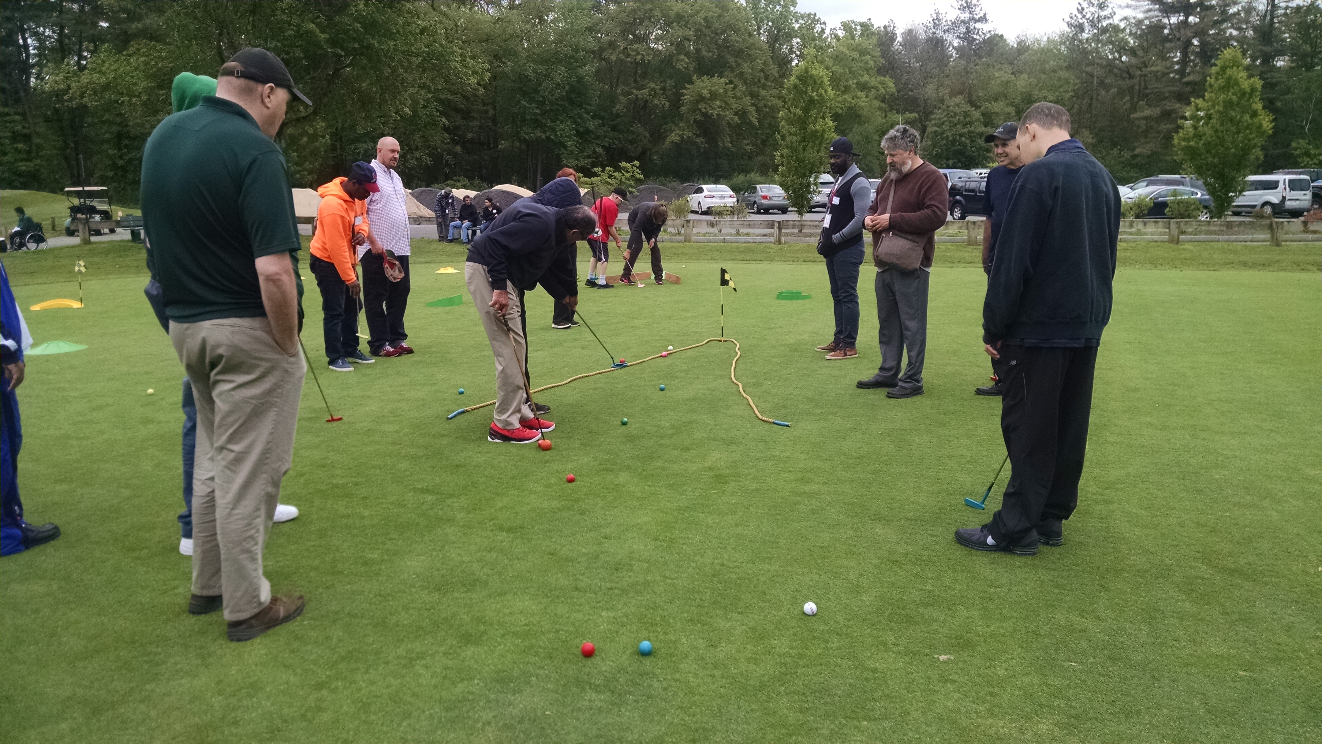 A group of about a dozen people is standing on a putting green. There is a rope on the green laid out in a V shape, with the hole at the tip of the V. Golfers are putting into the rope area using colorful golf clubs and golf balls.