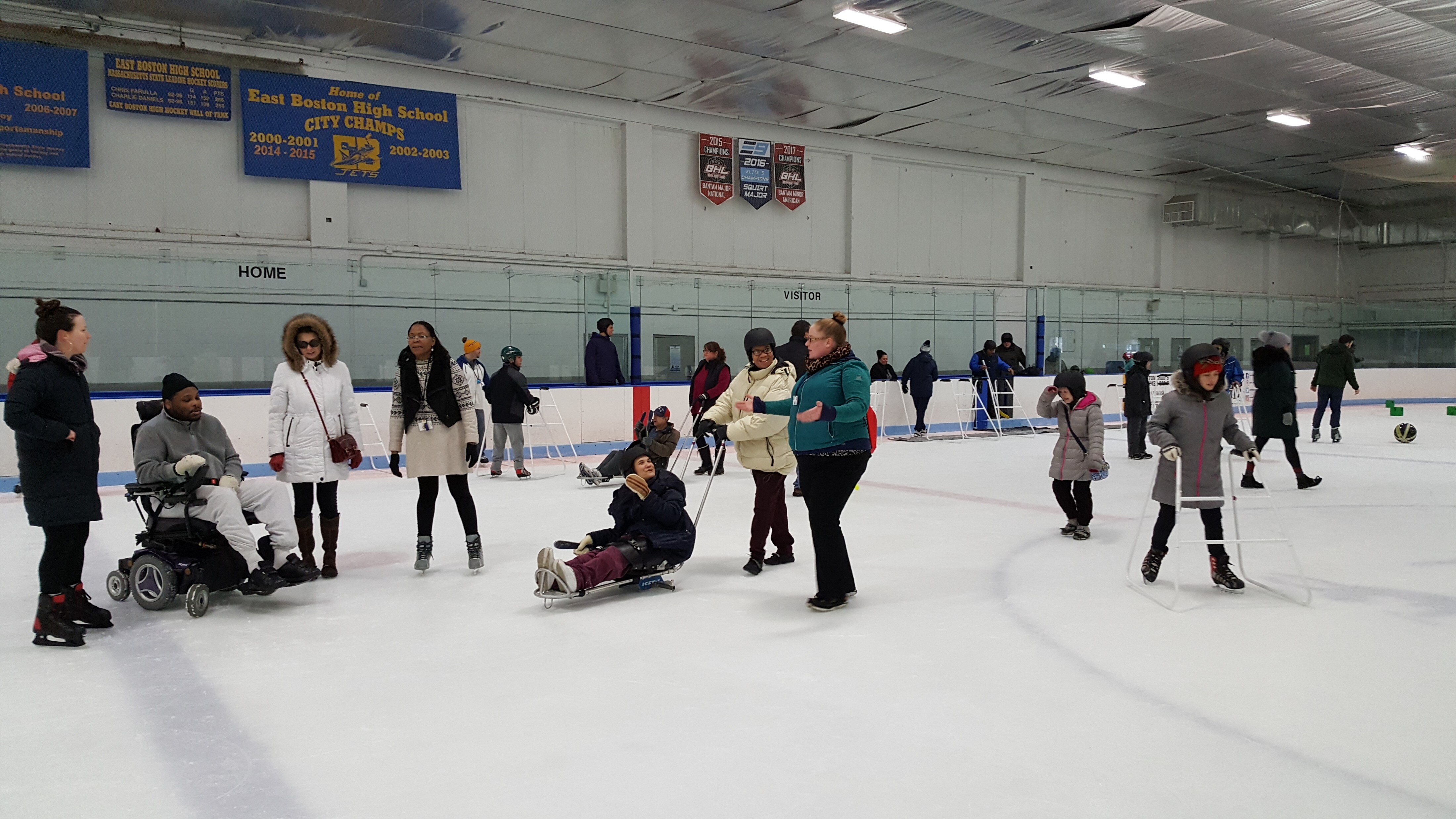 A large group of adults skating, walking and socializing in an ice skating rink wearing warm winter clothes. Some are on skating sleds, others using ice skates and walkers, some only using ice skates and one person is using a power wheelchair.