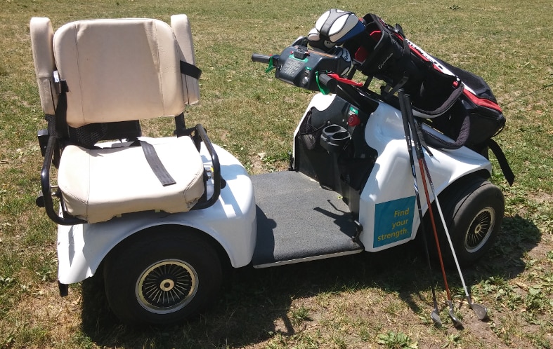 A SoloRider golf cart is sitting on the grass, with a bag of golf clubs resting in a rack on the hood. The cart has four wheels, and a flat smooth area between the front of the cart and the rear. The cart has two hand controls mounted on a column that extends from the hood. The seat is padded, with arm rests that swing up. The seat is turned so that it is facing 90 degrees away from the front of the cart.
