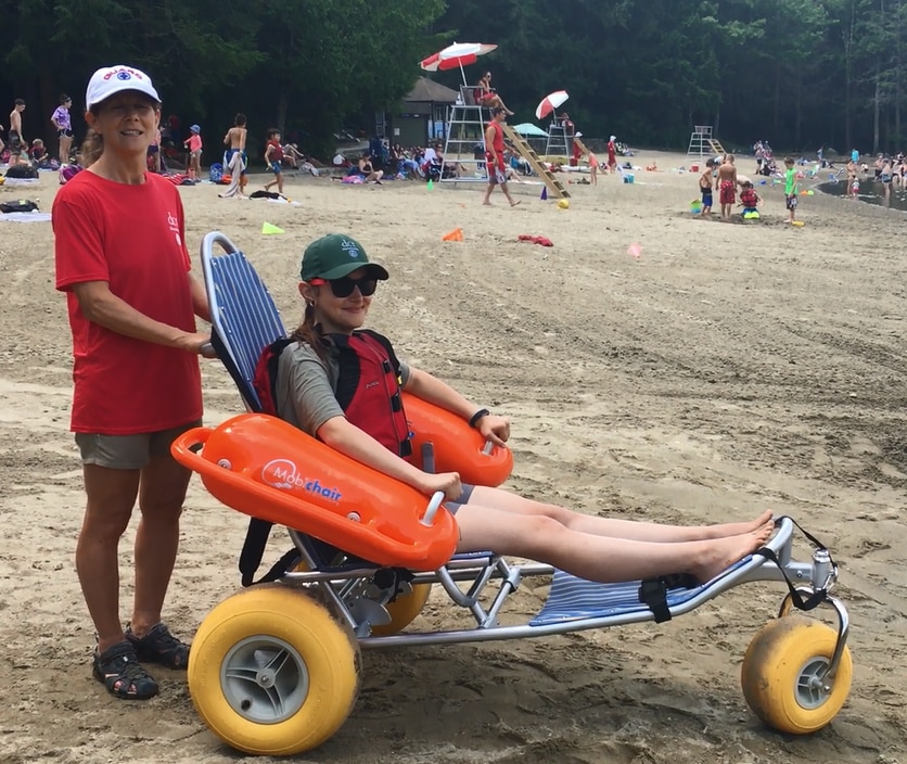 A woman is sitting in a beach wheelchair with a metal frame, fabric seat, and three balloon tires. The chair has large orange floats under the armrests. The woman's legs are stretched out in front of her and resting on the chair. She is wearing a personal flotation device around her chest. Another woman is standing behind her and holding onto the back of the chair.