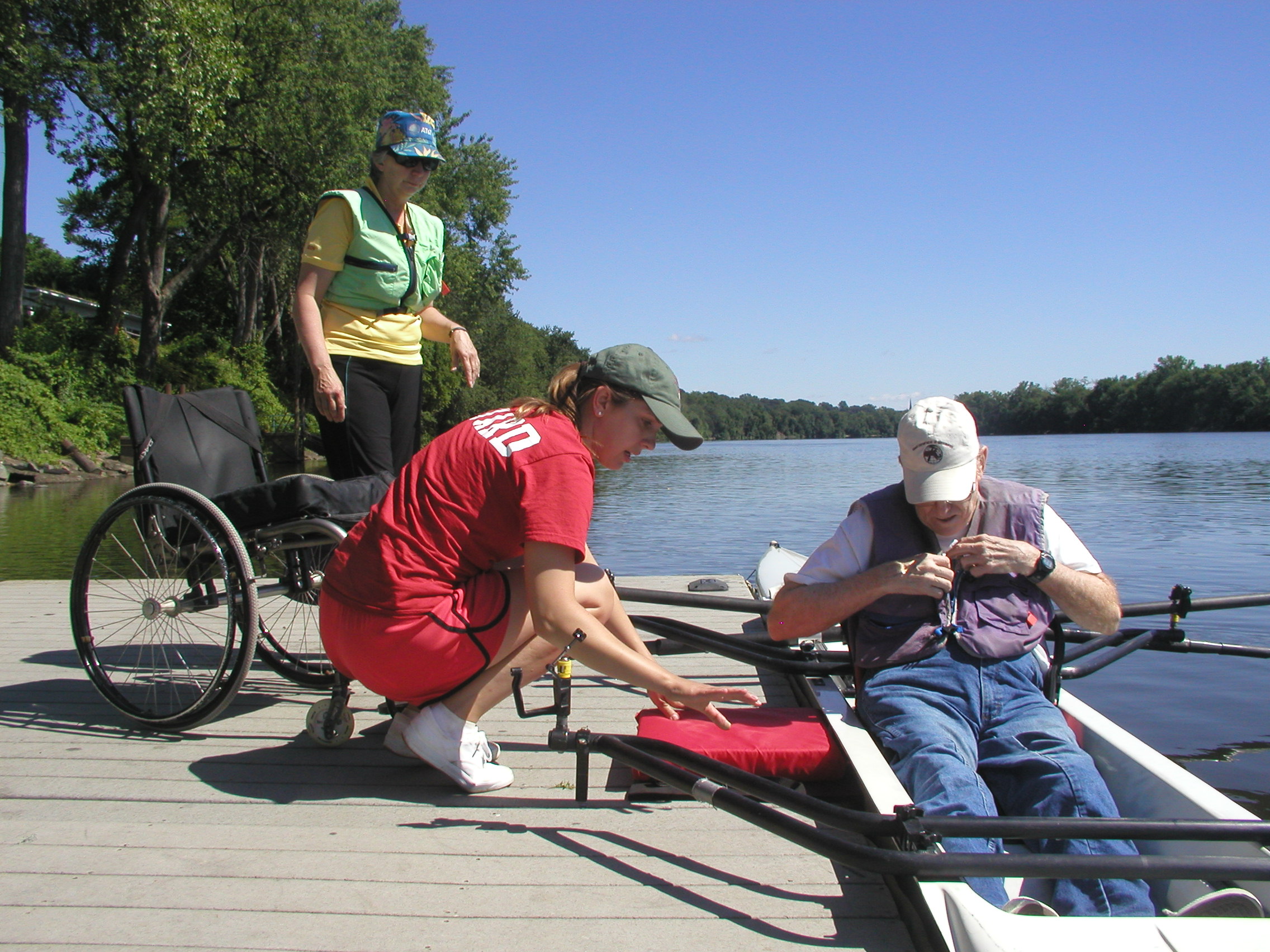 A rower is sitting in a shell in the water next to a dock. A lifeguard on the dock is adjusting a cushion placed on the dock next to the rower. Another person is standing next to a wheelchair on the dock.