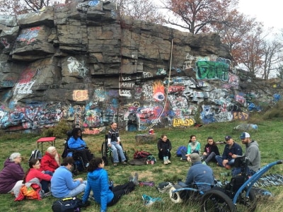 A group of people is sitting on grass in front of a rock face covered in graffiti. The rock face has climbing ropes hanging from it. Several of the people are using wheelchairs.