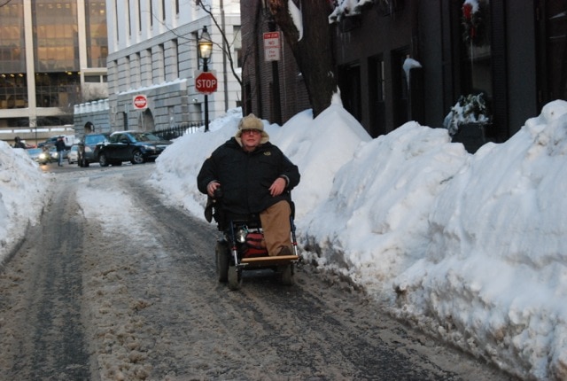 Tom, a white, middle-aged man, rides his wheelchair down a snowy downtown street. The street has been plowed and there are large snow banks on either side of him.