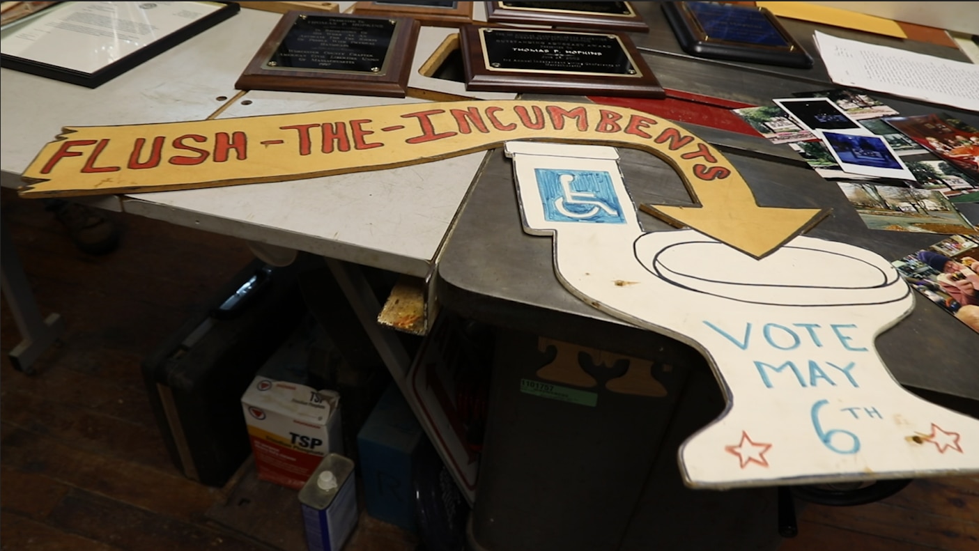 A handmade wooden sign in the shape of a toilet reads "Flush the Incumbents! Vote May 6th!" under the accessibility symbol