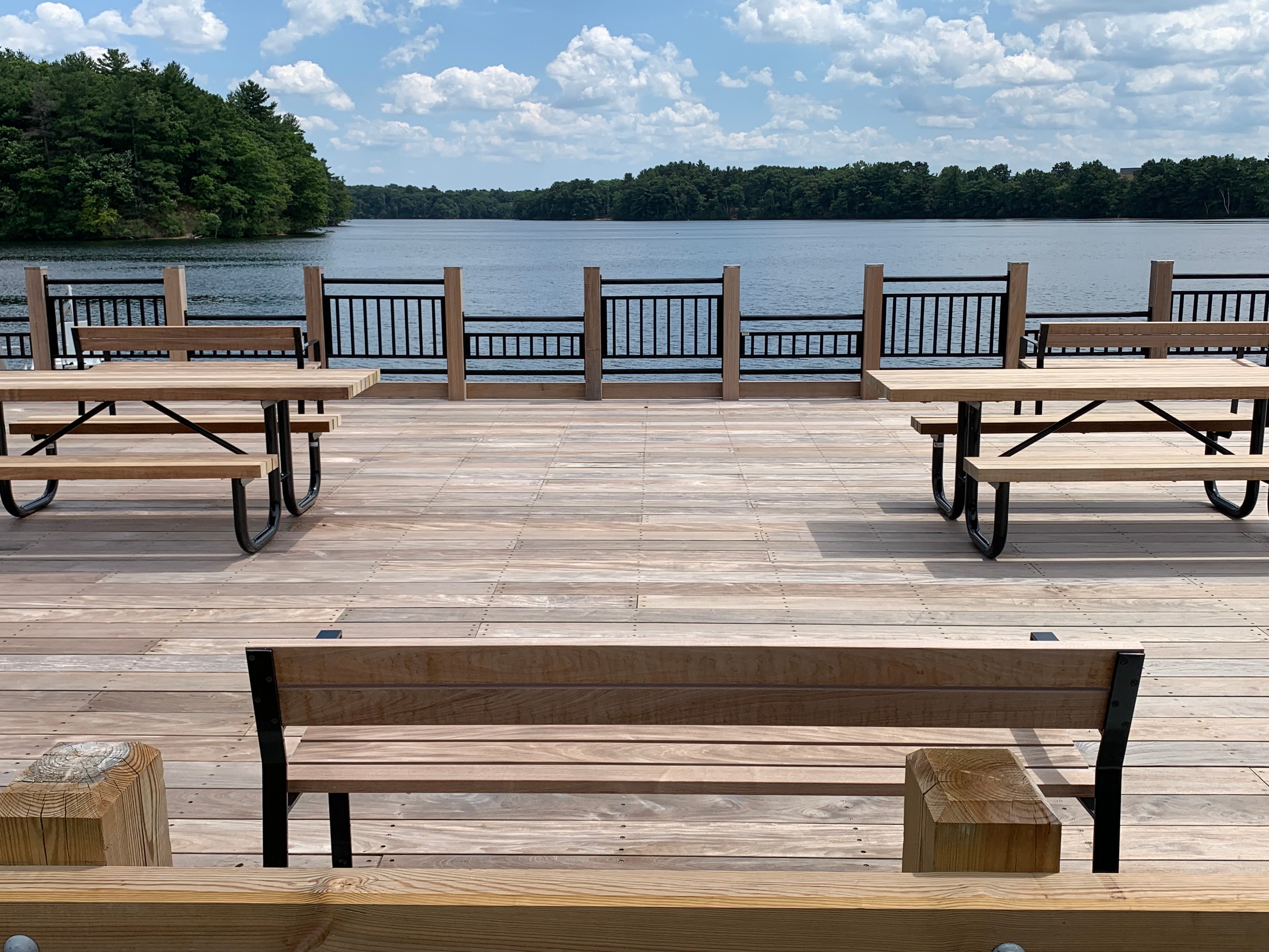 A large wooden deck looks out over a lake surrounded by trees. There are a bench and two accessible picnic tables on the deck. Every other section of the deck railing is lowered.