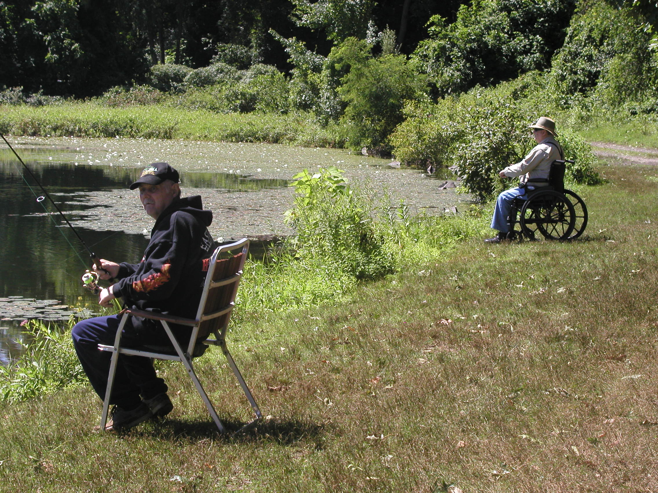 Two people are fishing on a grassy shoreline. One of the people is using a wheelchair.