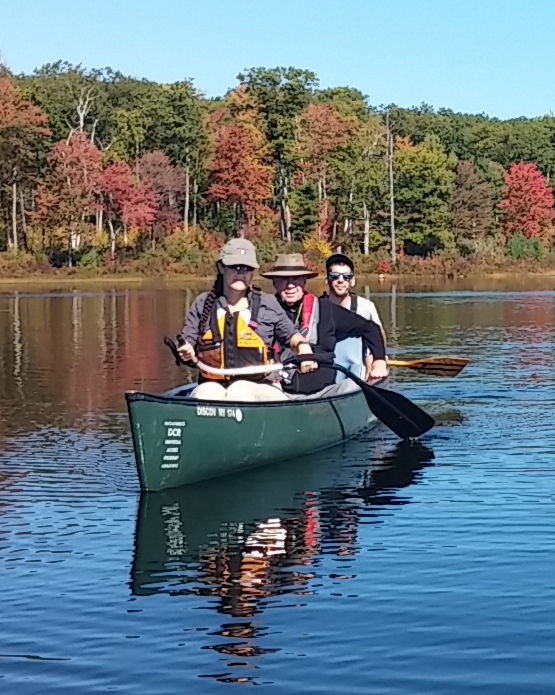 A paddler uses an S-shaped paddle. The end of the paddle curves up to the grip, the shaft lays across the lap, and the far end curves down so the paddle blade is in the water.