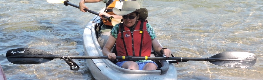 A paddler in the front of a tandem kayak is using paddle with attachments that jut off of it. One of the attachments is connected to a cuff on the paddler's wrist.