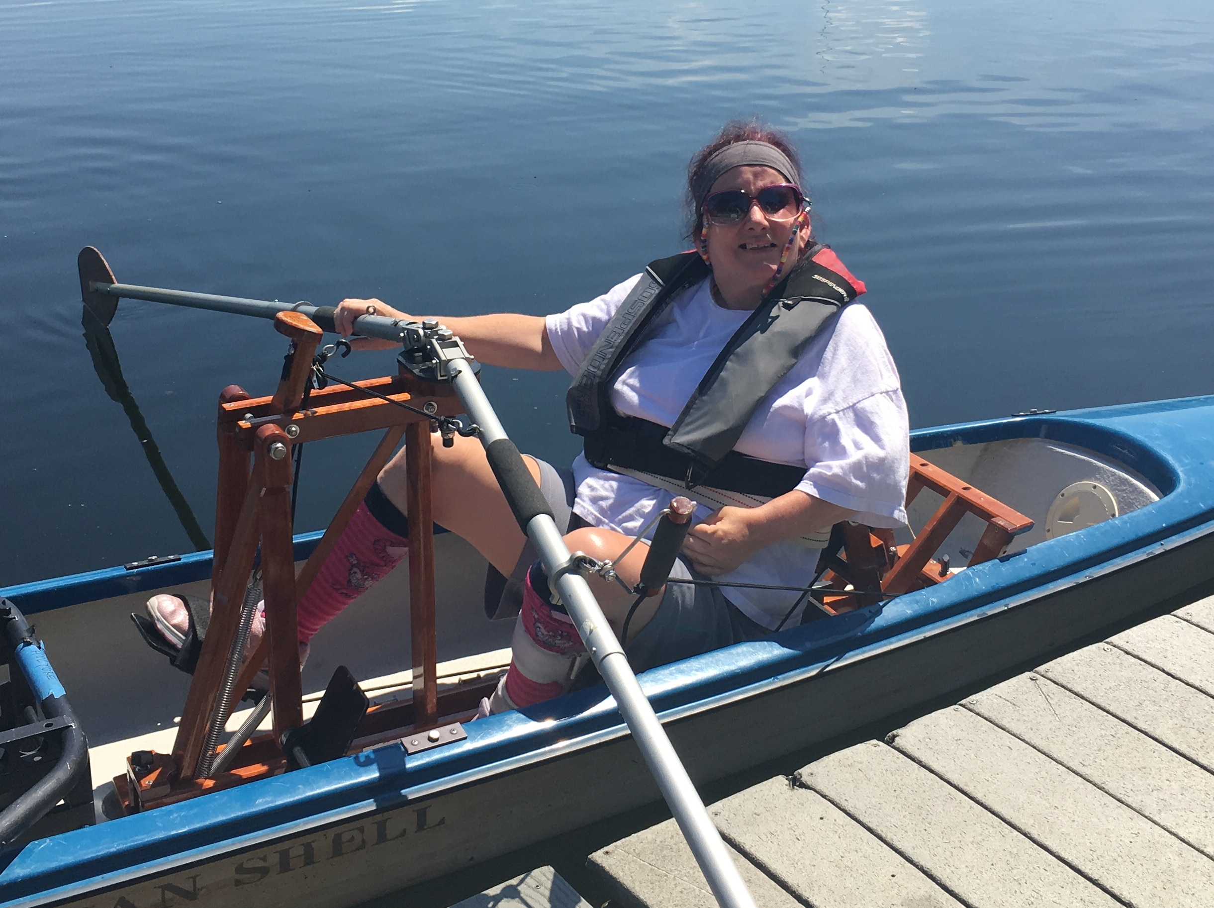 A person sits in a Frontrower in a shelll. The FrontRower is made of thin pieces of wood and raises the oars up to chest height. Footpedals attach to an arm that can move the oars.