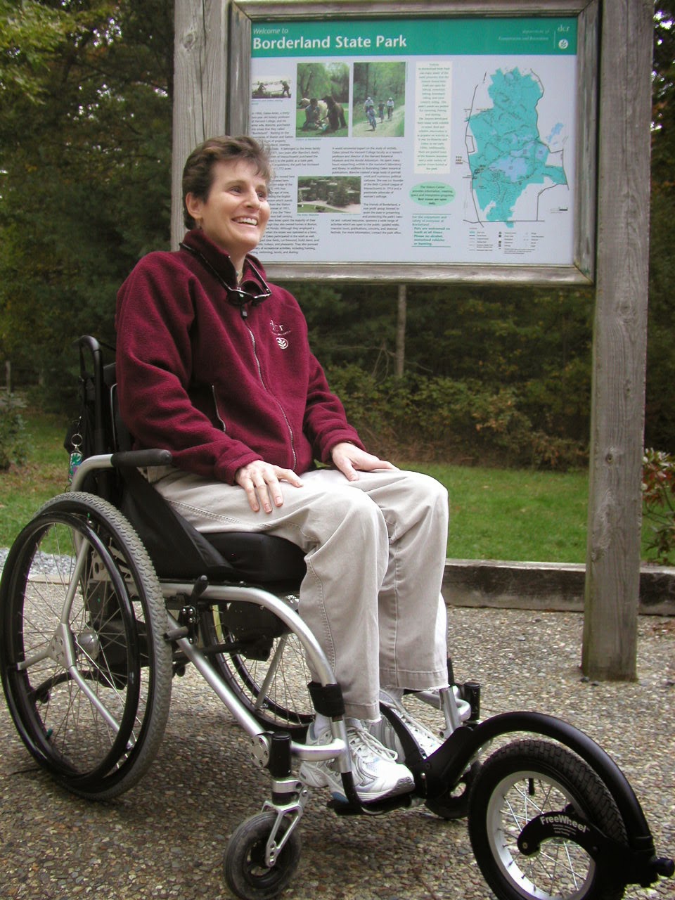 A woman sits in a wheelchair with a Freewheel attached on the front. A kiosk behind her reads Borderland State Park.