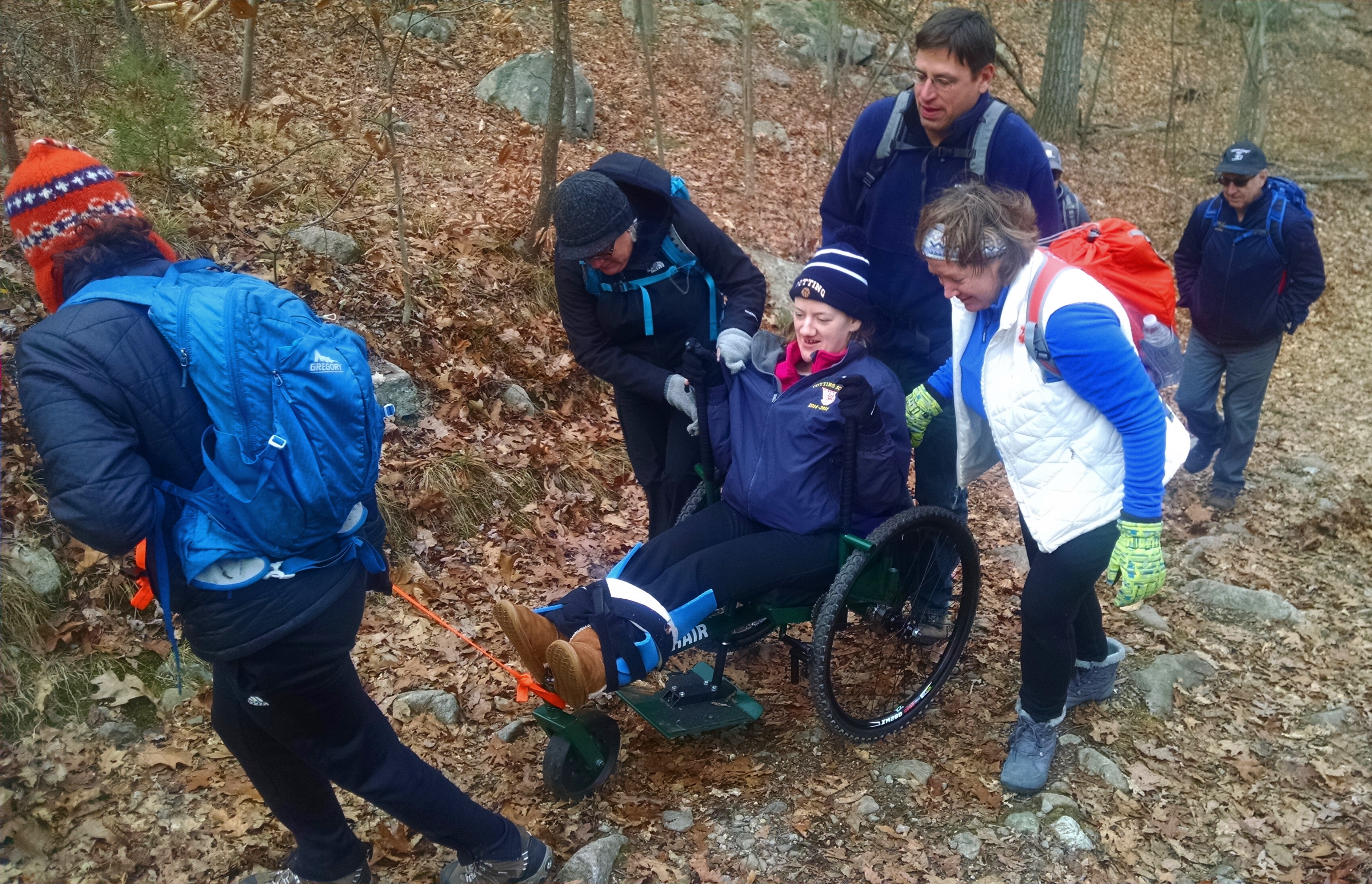 A hiker in a GRIT Freedom Chair is being pushed up a hilly trail by several hikers behind the chair. A strap on the front of the chair is being used to pull it as well.