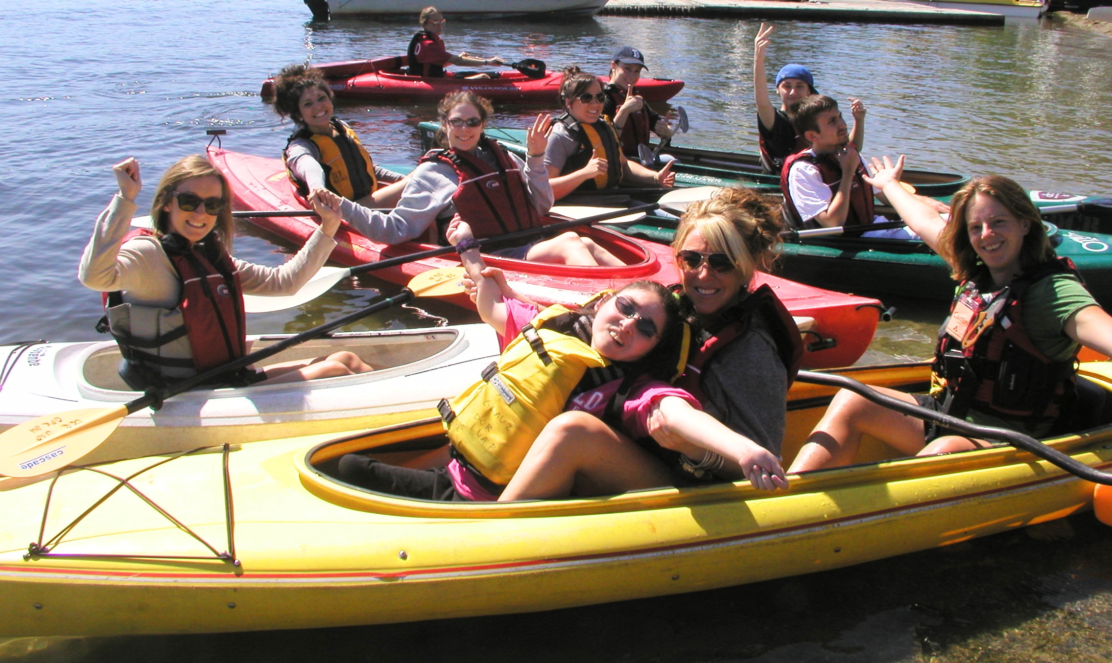 A mix of single and tandem kayaks on the water. The paddlers are raising their hands in the air and smiling. One paddler is sitting in the lap of another paddler.