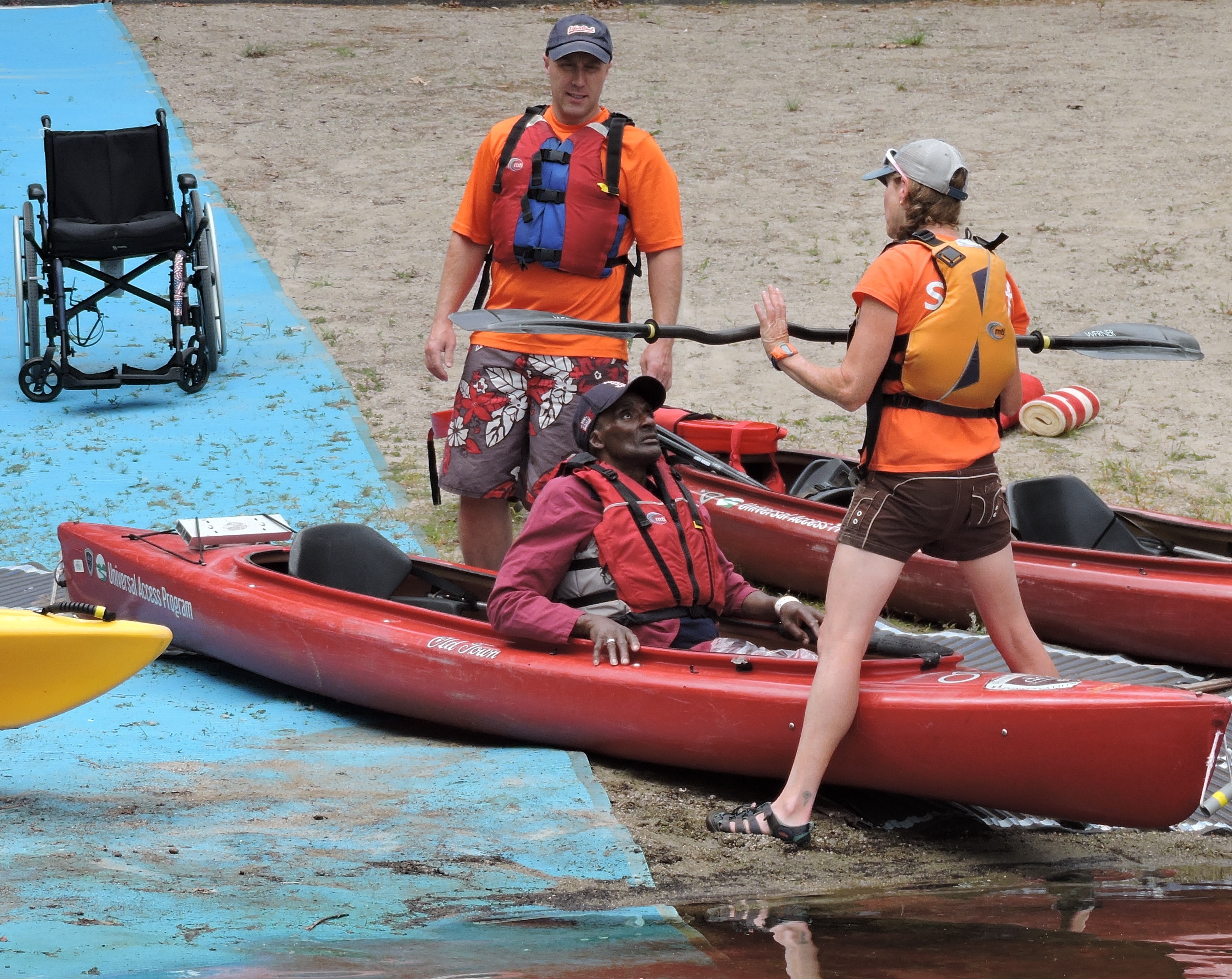 One paddler is sitting in a tandem kayak while another stands on the beach. An instructor is straddling the kayak and holding up a paddle. A beach mat with a wheelchair on it leads down to the kayak.