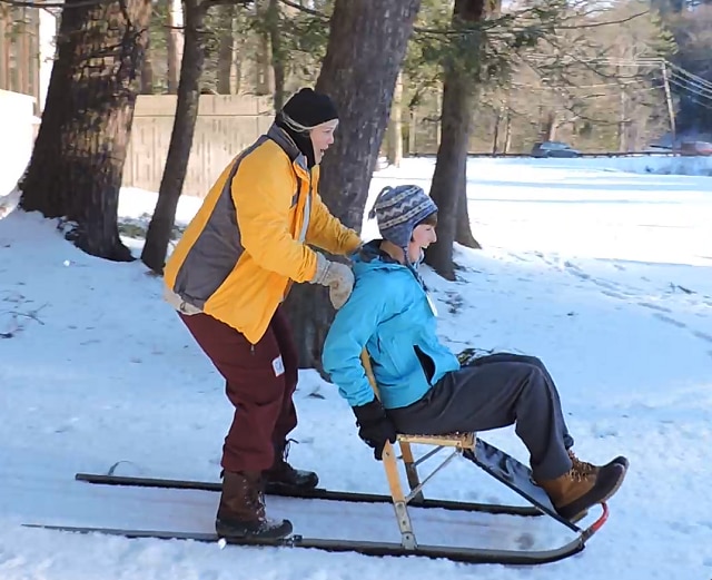 Two people are using a kicksled. The person on the back is standing on the runners, while the person in front sits in the chair. They are sledding down a hill and laughing.