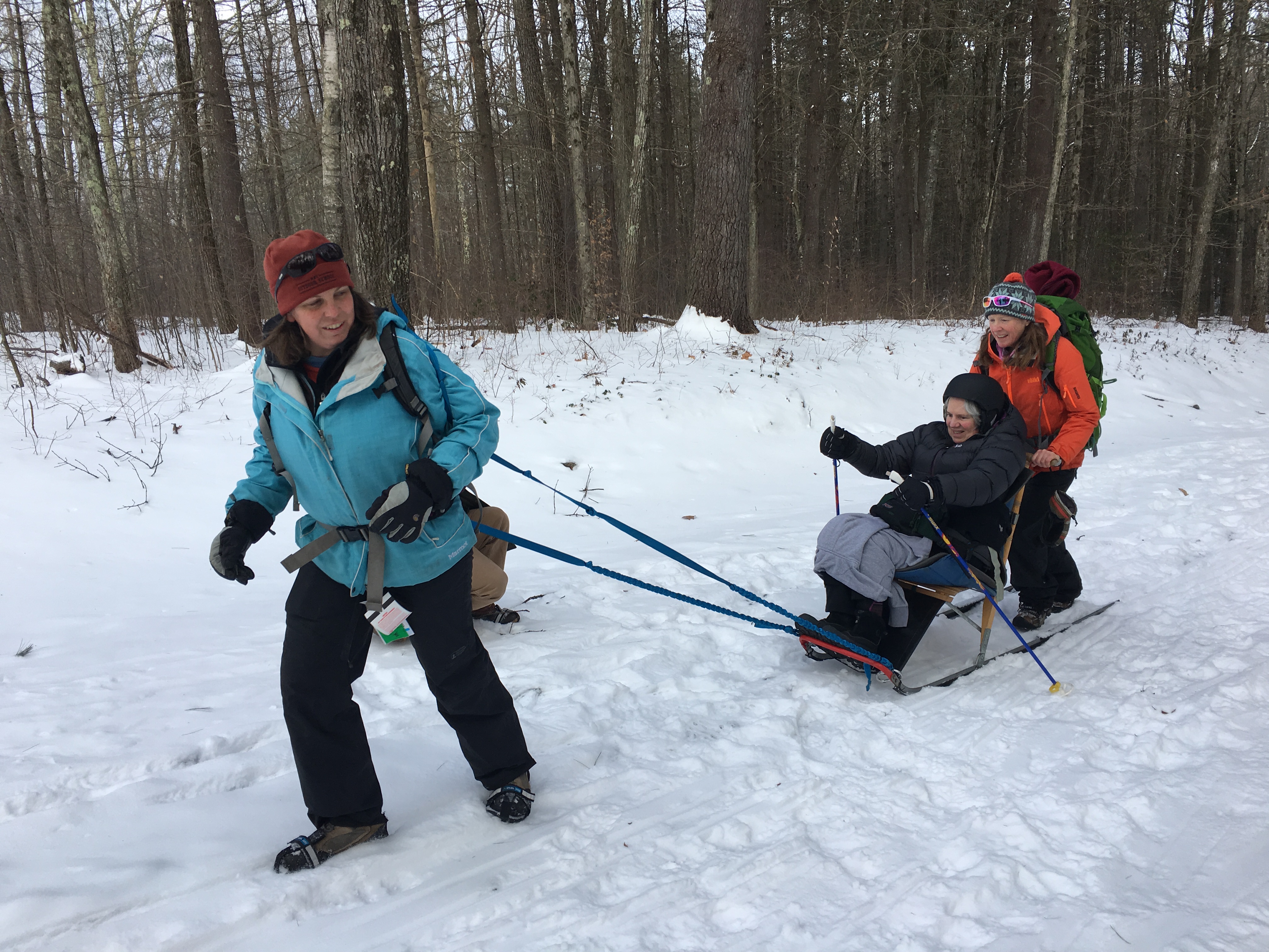 A skier in a kick sled is using poles to push it up a slight hill. Another person is standing behind the kick sled and pushing, while a third person tows the sled with a strap.