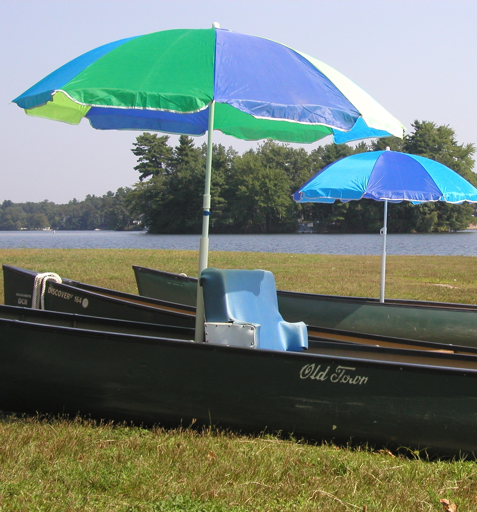 A padded seat sits in the bottom of a canoe on the shore. An umbrella is mounted over the seat.