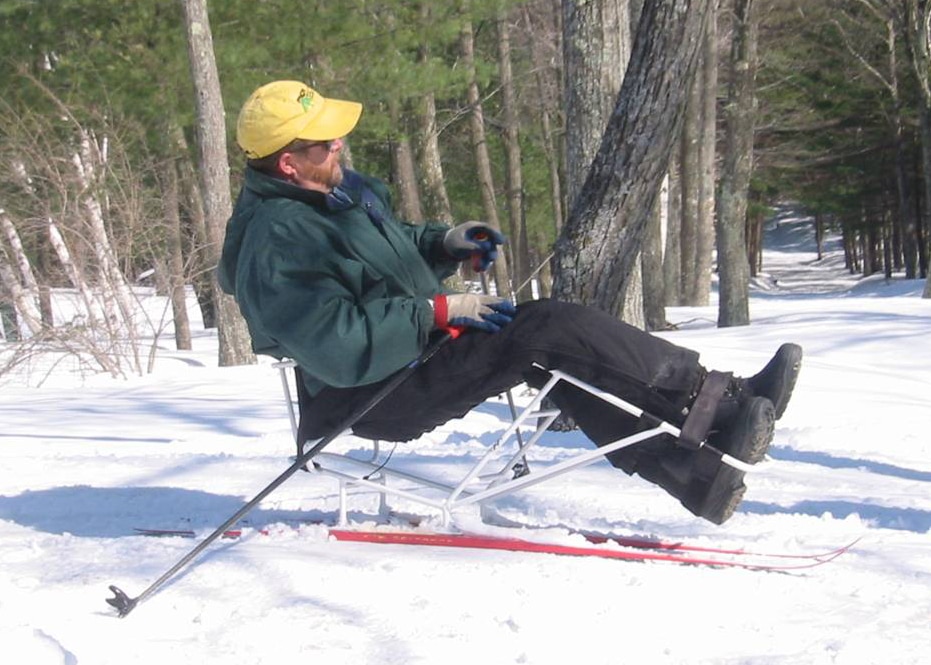 A skier is sitting in a sit ski with frame that bends up at the knees. The skier's kegs are strapped into the ski frame. The skier is using two short ski poles to push himself through the snow.