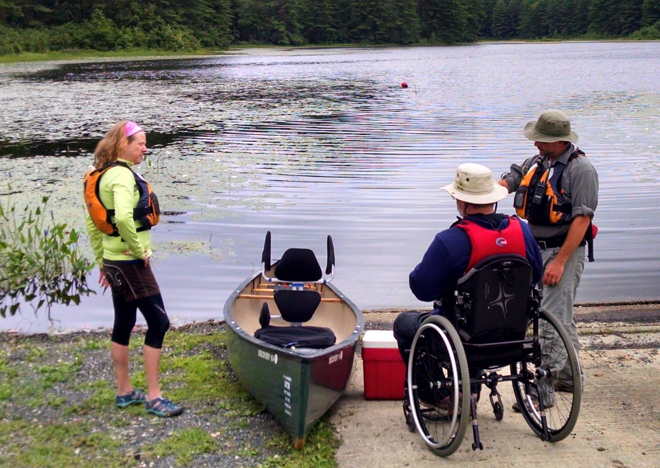 A paddler in a manual wheelchair sits next to a canoe with an adaptive seat. A cooler is next to the canoe and two program staff are nearby.