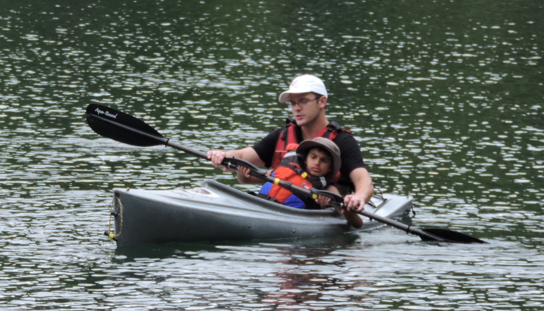 A child sits in the lap of an adult in a kayak. They are both holding a paddle and paddling together.