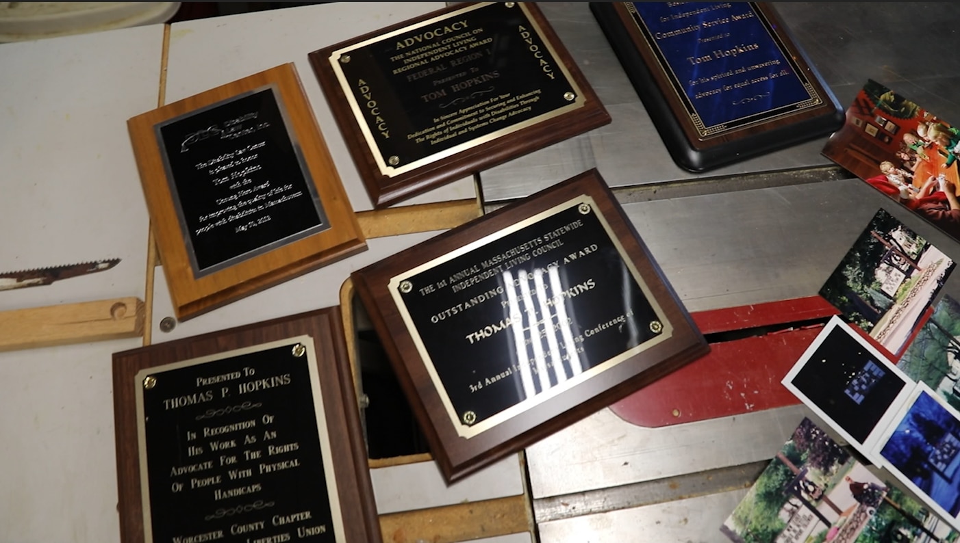 Several of Tom's advocacy awards are laid out on a table in his woodshop. Much of the writing is blurry, but an award from the Massachusetts Statewide Independent Living Council is front and center.