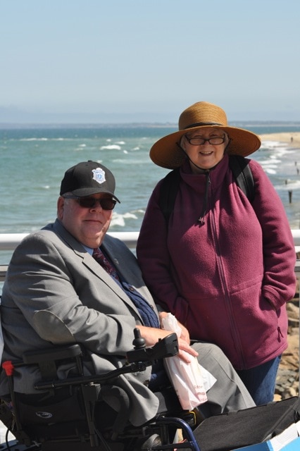 Tom and his wife, Linda, at the beach. Tom is wearing a gray suit and a black baseball cap and Linda stands next to him in a sun hat and a maroon fleece jacket.