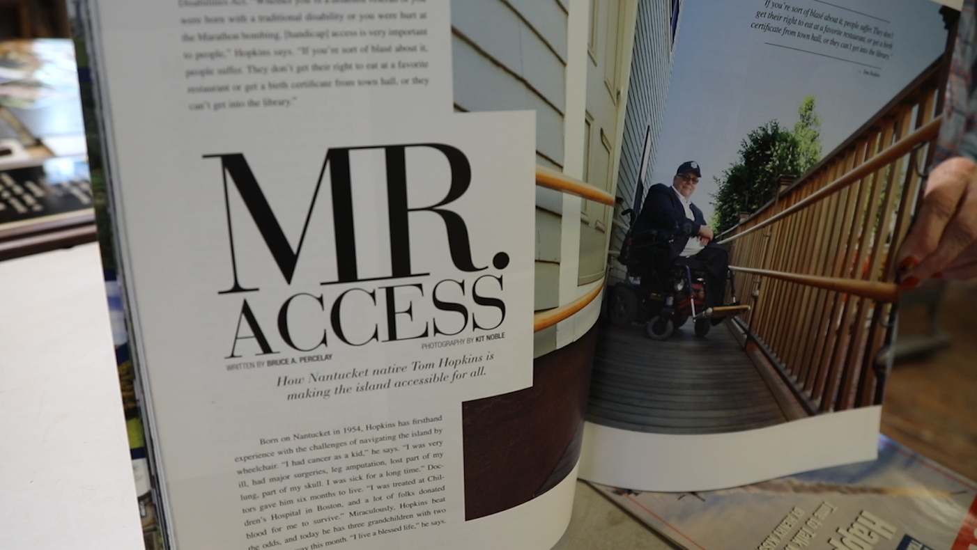 A magazine is opened to a page with the title "Mr. Access: How Nantucket native Tom Hopkins is making the island accessible for all" and a photo of Tom wearing a navy blue suit on a wooden boardwalk.