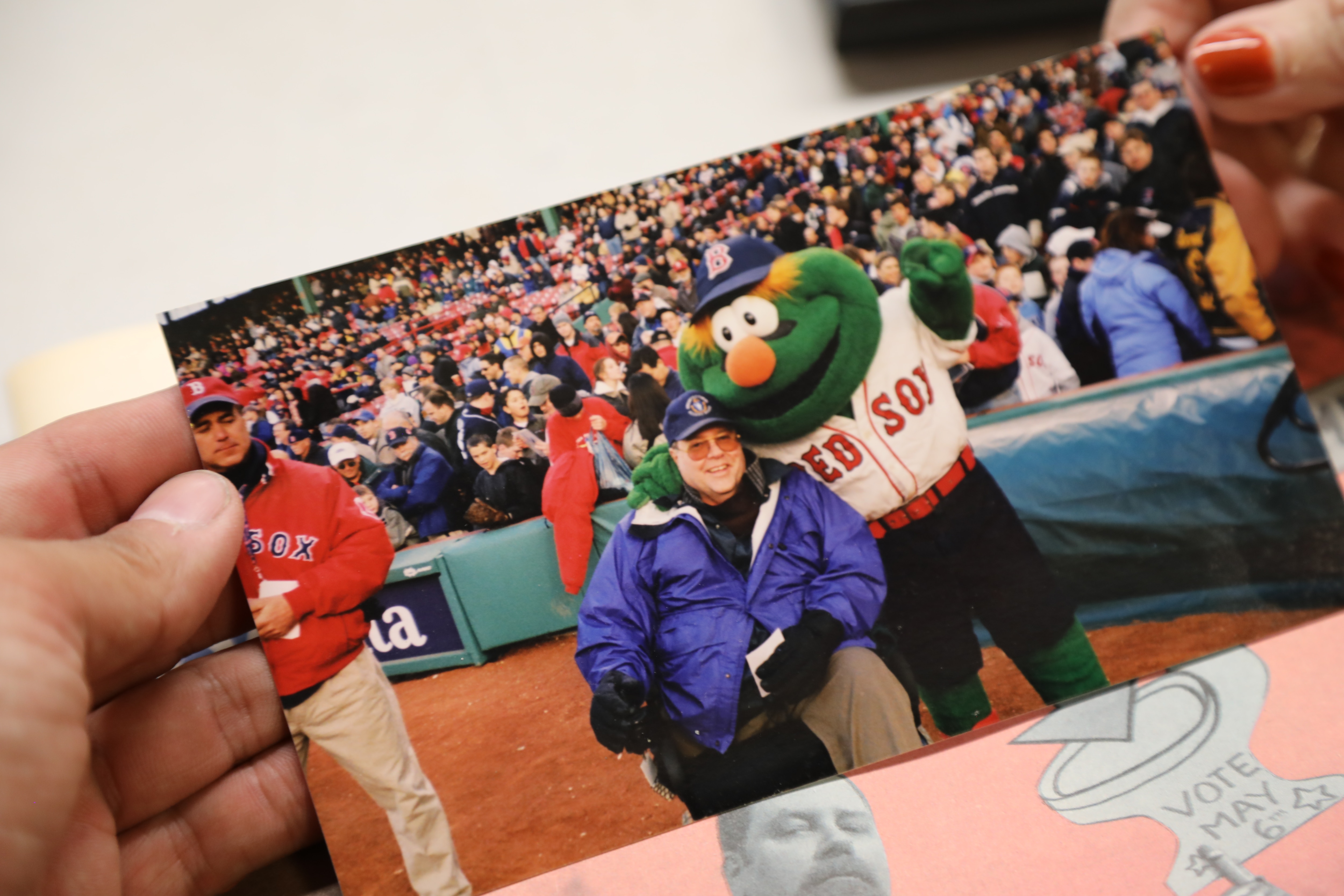 Tom poses with Wally the Green Monster at a Red Sox game. They are on the field and the stands are full behind them