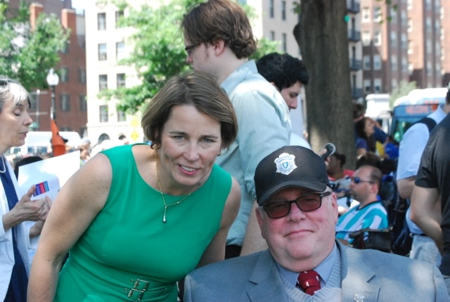 Tom poses outdoors on a busy street with Maura Healey. He is wearing a suit and a baseball cap with a badge on it