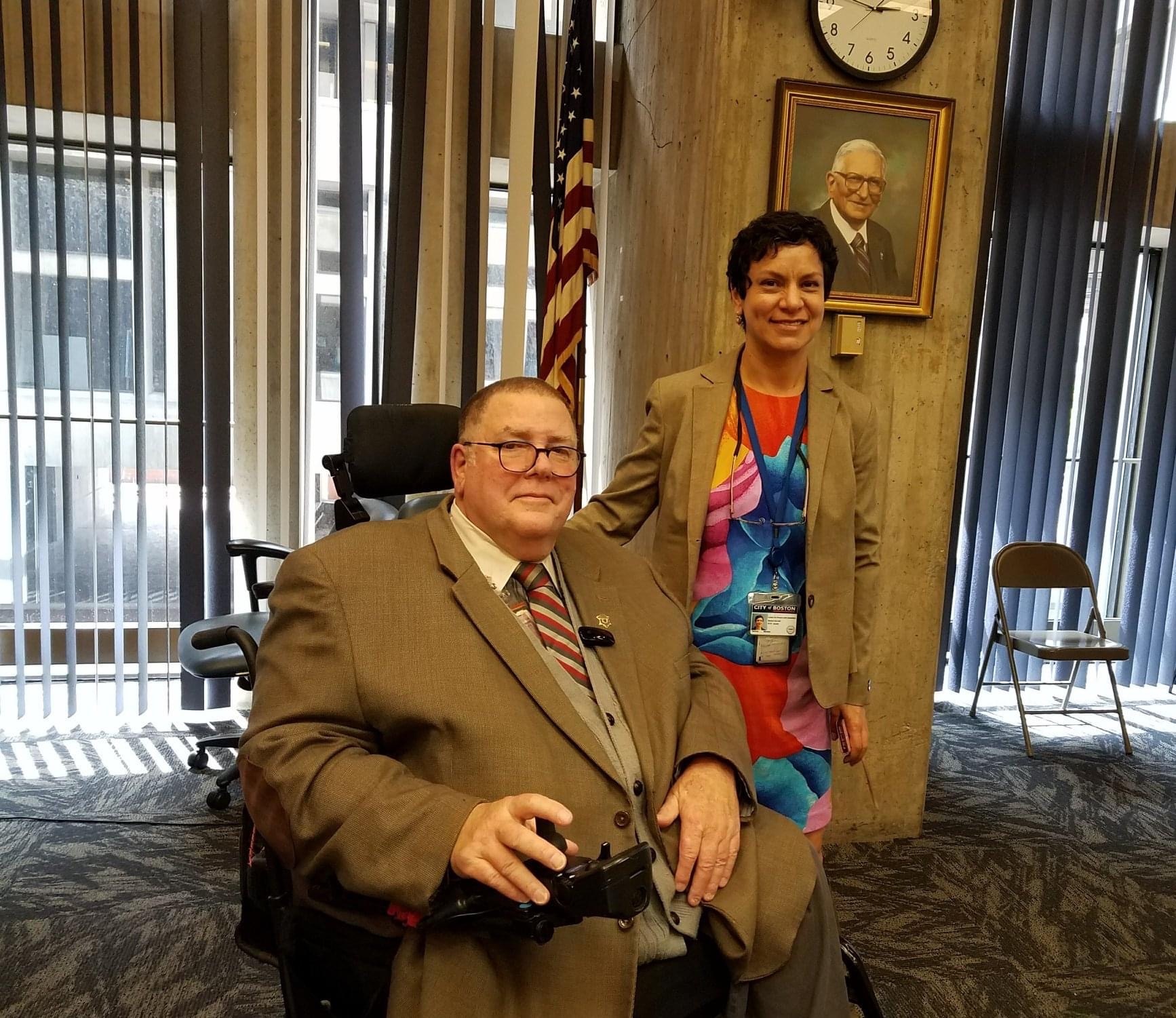 Tom, wearing a beige suit, poses next to Patricia Mendez. They are in an office with the American flag behind them and a portrait hanging on the wall.