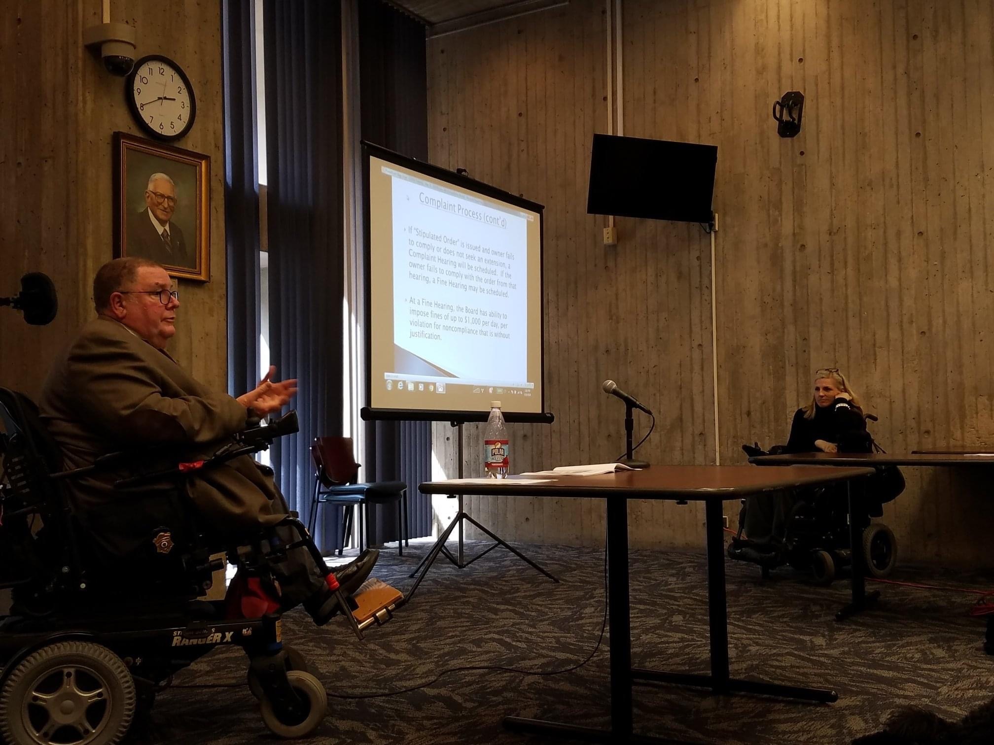 Tom sits at a table with a microphone presenting in an office setting. A large screen is to his left with a PowerPoint slide visible on it. A woman is seen sitting in a wheelchair in the distance.
