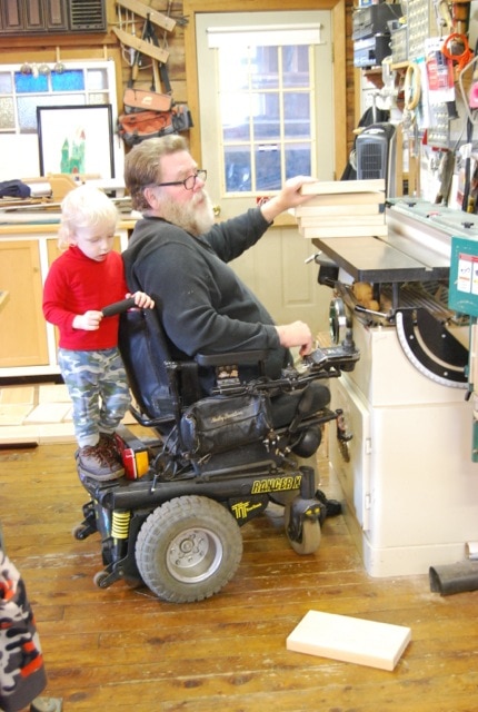 Tom sits at a work table in his wood shop. His granddaughter stands on the back of his wheelchair gripping the handle.