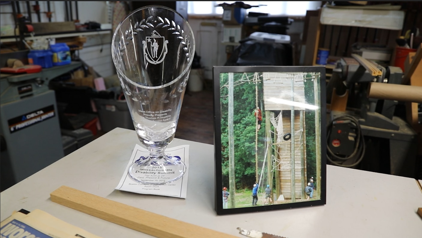 Tom's 2015 Thomas P. Hopkins award, a crystal vase etched with the Massachusetts state seal, sits on a table in Tom's woodshop next to a framed photo of Tom climbing up a tall wooden beam at a high ropes course