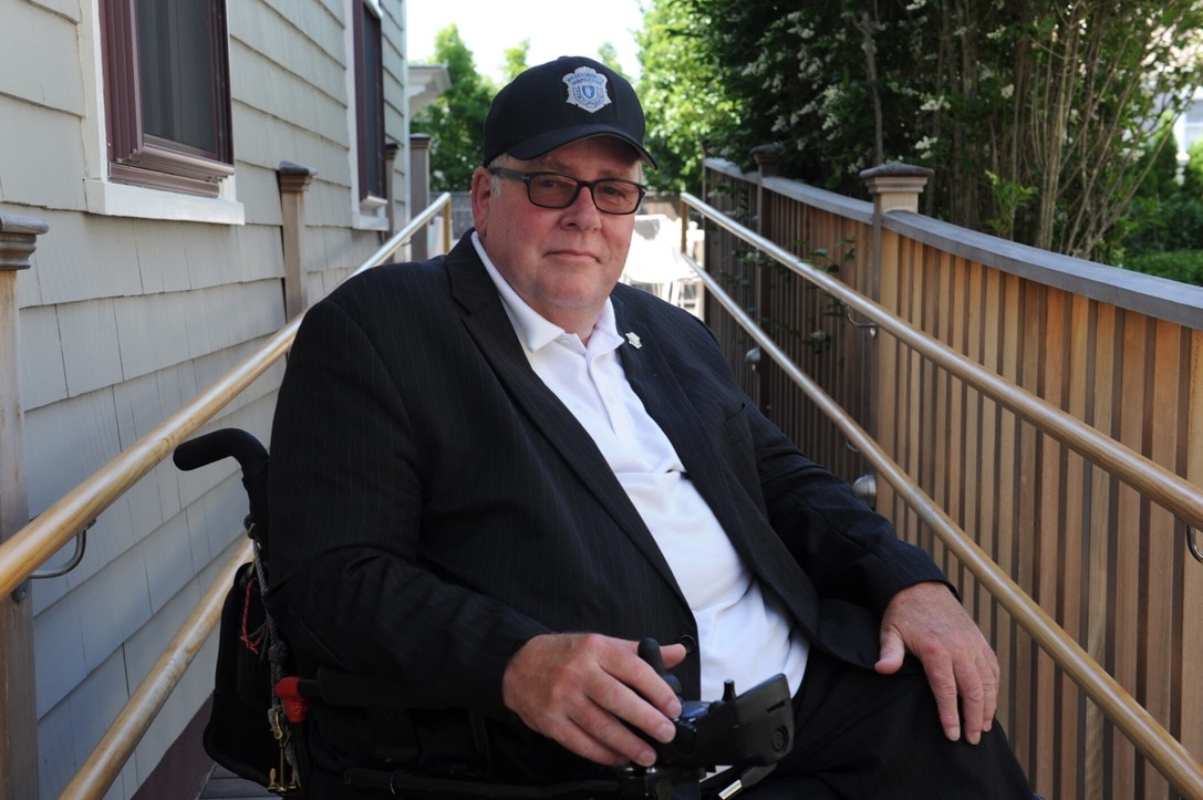 Tom in middle age, sitting in his wheelchair at the end of a wooden ramp with handrails on either side. He is wearing a black suit and black baseball cap with a badge on it.