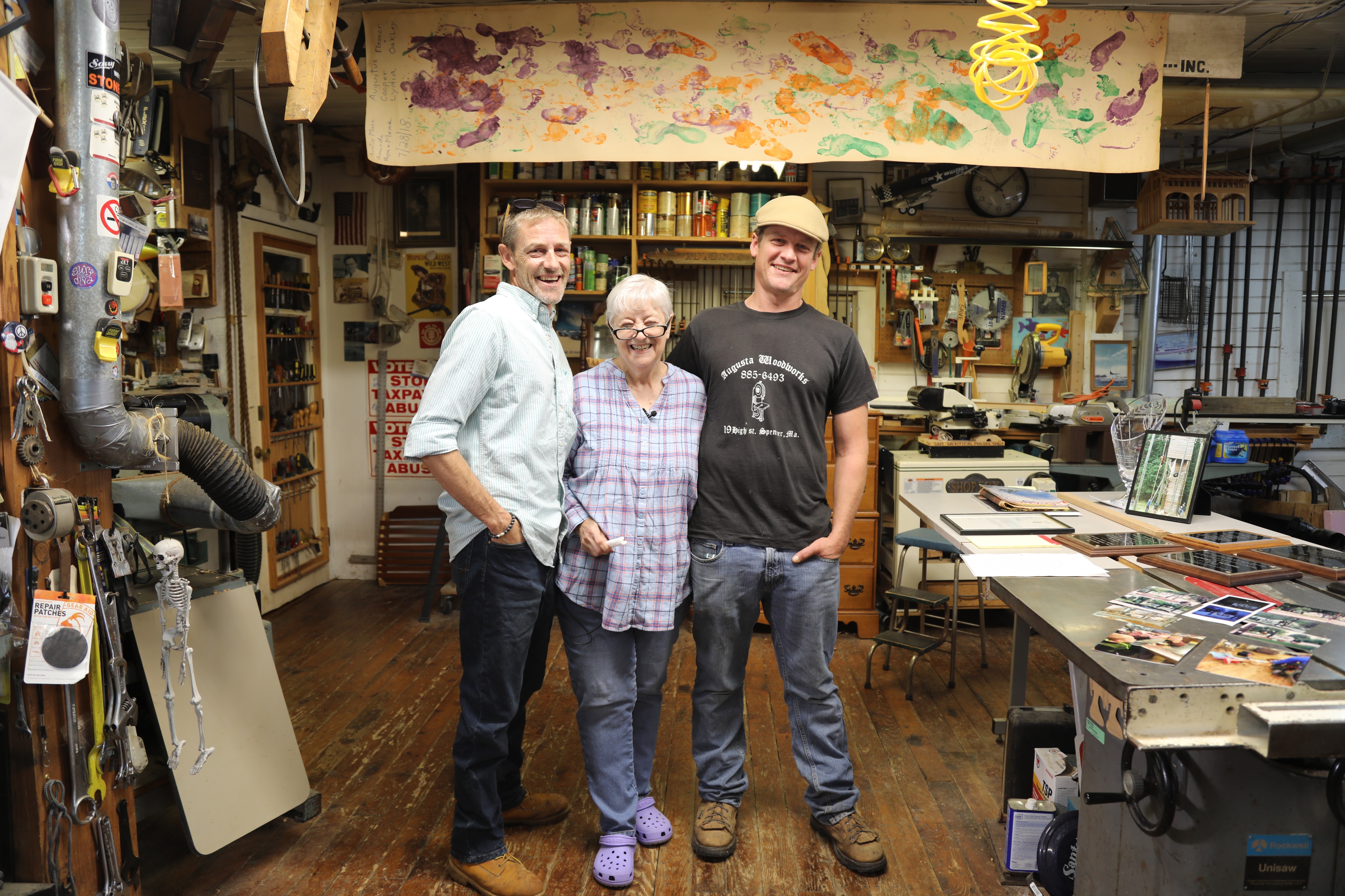 From left to right, Damon, Linda, and Justin Hopkins stand posing and smiling in Tom's woodshop. A paper banner hangs above them with footprints and handprints in orange, purple, and green paint. The woodshop is cluttered with Tom's awards and tools.