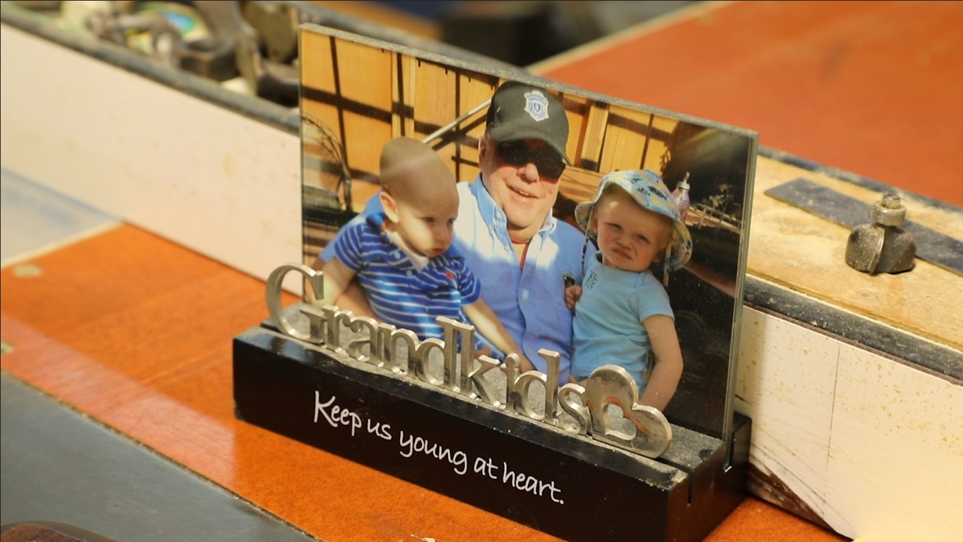 A small framed photo of Tom with his two grandchildren. His grandson, a baby, sits on his lap on his right side and his granddaughter, a toddler, sits on his lap on his left side. They are outside on a sunny day. The frame reads "Grandkids keep us young at heart."
