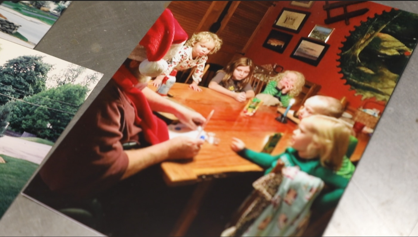 Tom, his back to the camera, sits at a wooden dining table with a Santa hat on. His five grandchildren, all toddlers and young children, sit around the table with him. They are all wearing pajamas and looking intently at a paper Tom is holding in his hands.