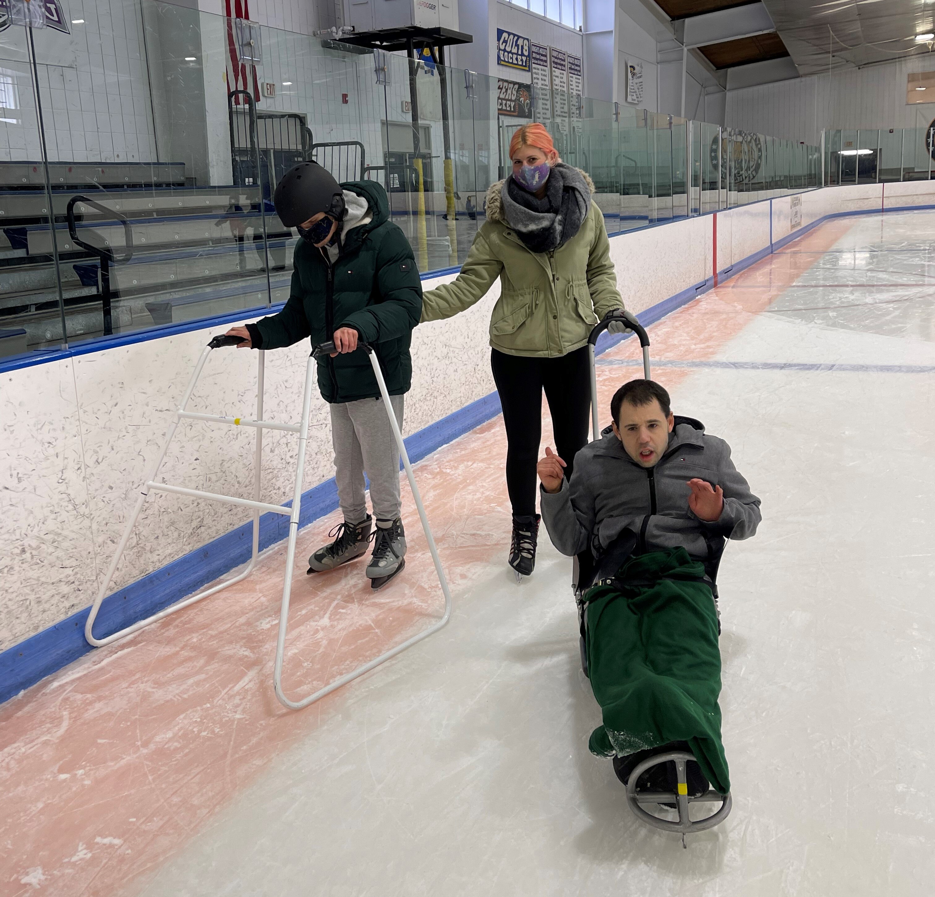 Three people ice skating at an indoor rink. One is on ice skates and pushing a sled. Another skating on a ice sled looking at the camera. The third ice skating using conventional skates and a walker. 