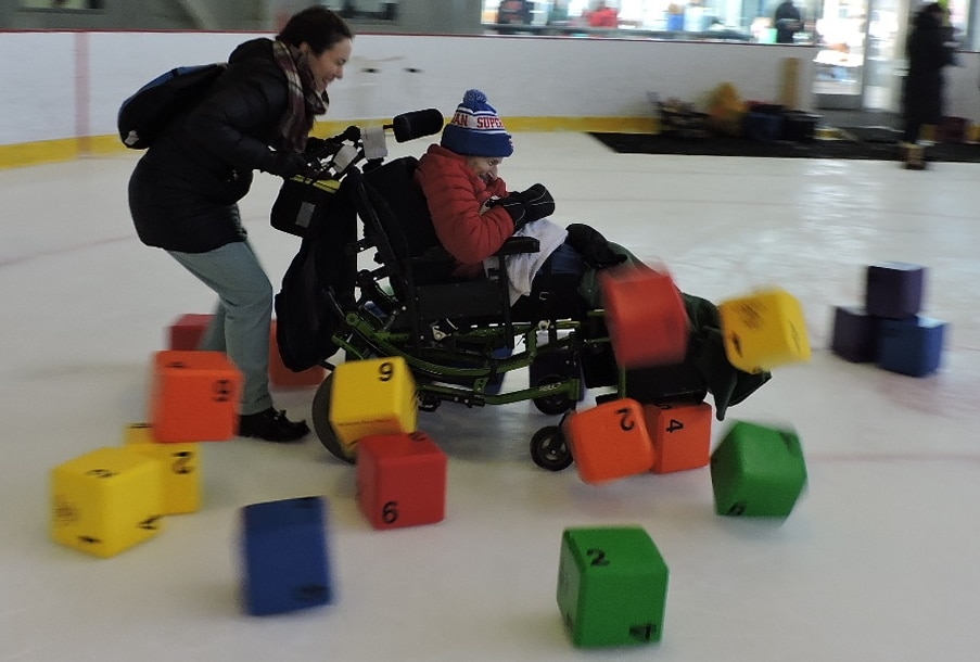 A skater in a wheelchair is crashing through a tower of colorful blocks while being pushed.